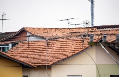 Concerned Homeowner Stands In Front Of Their House, Looking Up At A Roof Damaged By A Recent Storm Under A Dramatic, Overc...