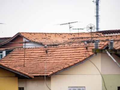 Concerned Homeowner Stands In Front Of Their House, Looking Up At A Roof Damaged By A Recent Storm Under A Dramatic, Overc...