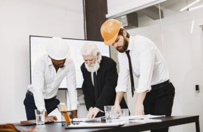 Diverse Team Of Professionals In A Modern, Bright Office Collaborating Over Architectural Blueprints Spread Out On A Large...