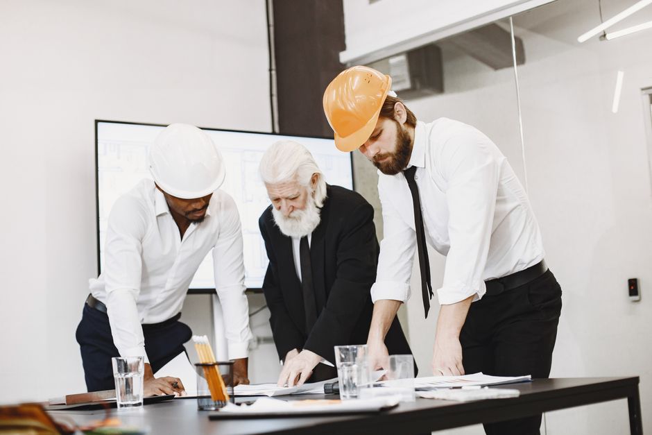 Diverse team of professionals in a modern, bright office collaborating over architectural blueprints spread out on a large...