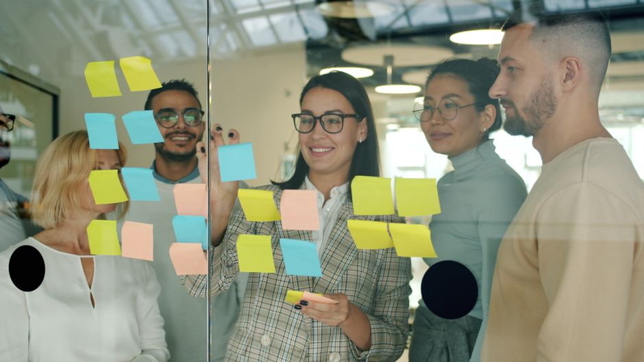 A dynamic group of colleagues brainstorming with colorful sticky notes and markers on a large glass wall in a modern workspace.